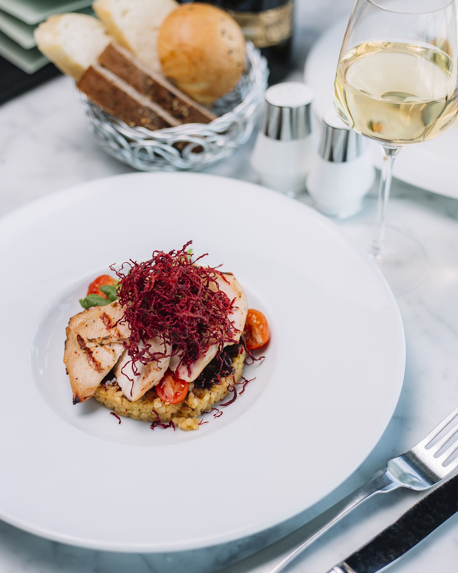 ブルグルと野菜の上に盛り付けられたグリルチキンフィレ、赤キャベツのフライを添えた料理。白ワインとフレッシュなパンのバスケットが横に置かれている。